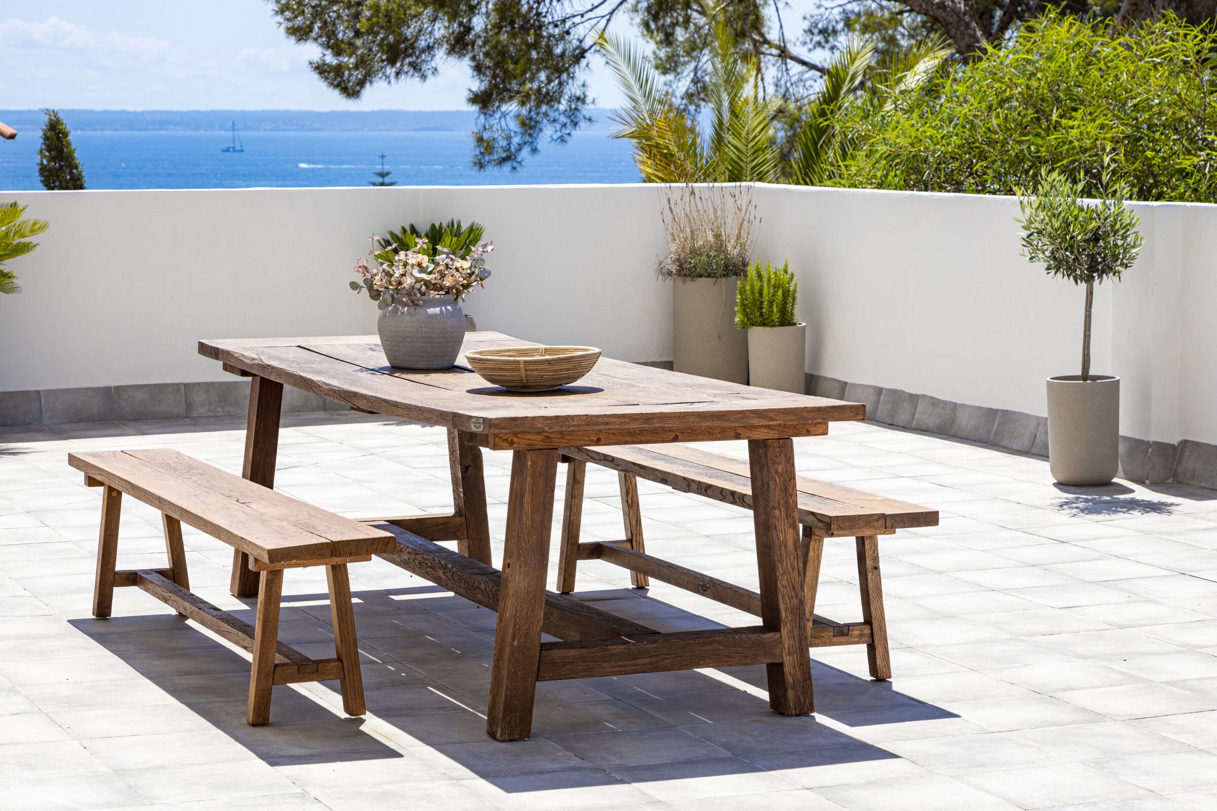 Outdoor terrace dining table of a home in Mallorca developed by Idaforma, framed by Mediterranean pines with the bay beyond.